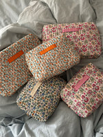 Fem quiltede makeup-tasker med blomsterprint, hver med et "LA PETITE CACHOTIÈRE" -label, ligger oven på et lyseblåt sengetæppe.