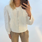 En kvinde tager en selfie med sin telefon. Hun er iført en hvid bluse med broderi og brune bukser, stående foran en hvid baggrund.