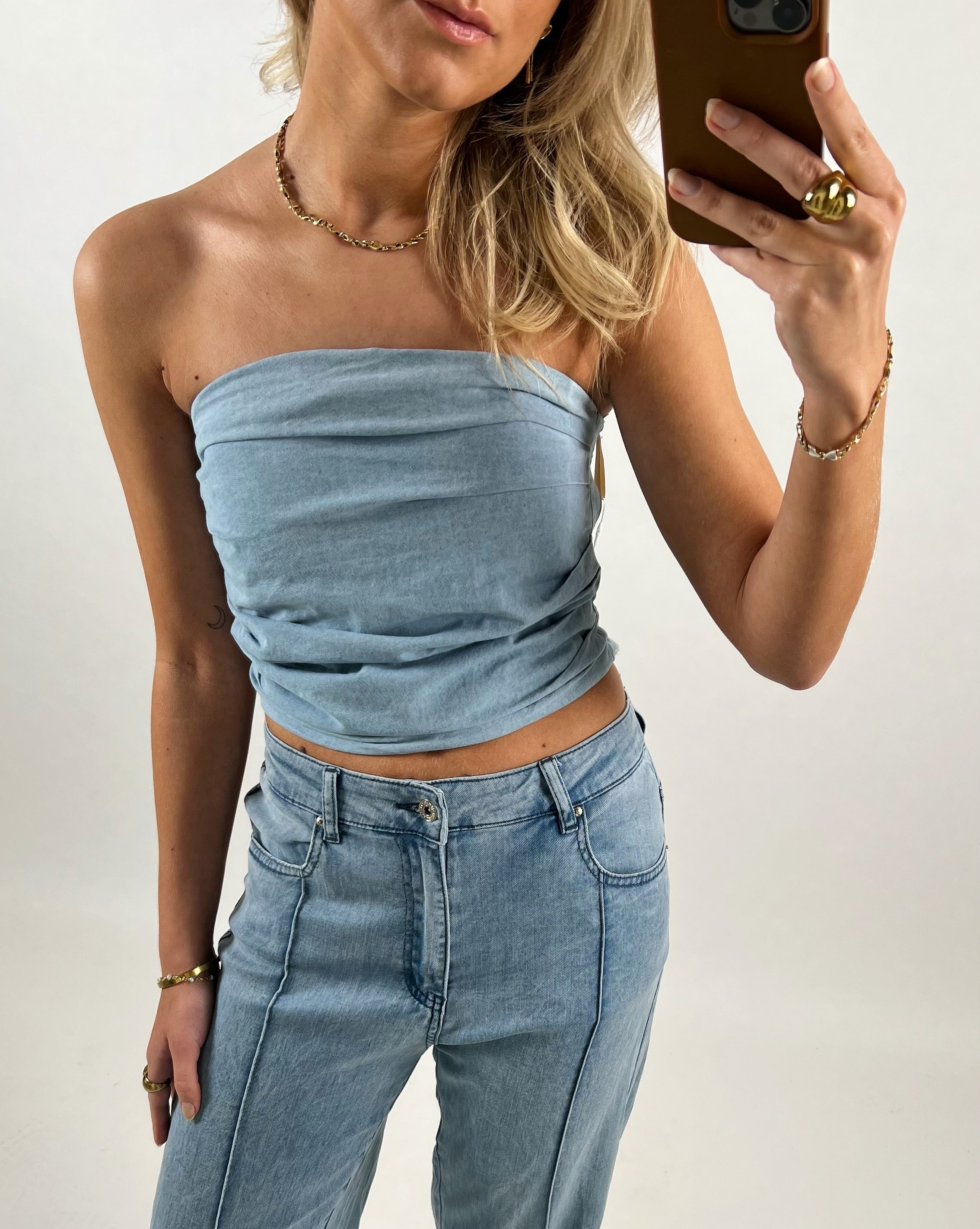 En kvinde iført en stropløs, lyseblå top og jeans tager et spejlfoto med en telefon. Hun bærer guldsmykker. Baggrunden er en neutral, hvid væg.