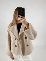 En person iført en beige frakke tager et billede med en sort telefon. Personen står i et neutralt, lysegråt rum.