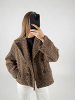 Kvinde iført brun, lodden jakke tager et selfie med sort telefon. Hun står i et lyst rum og har en sort trøje og hvide bukser på.