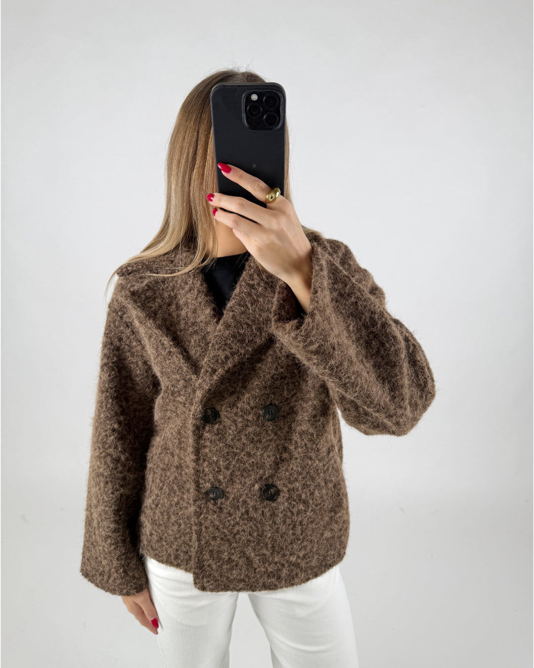 En person iført en brun, uldet jakke holder en sort smartphone op foran ansigtet. Personen står i et minimalistisk rum med en hvid baggrund.