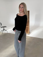 En person står afslappet iført en sort trøje og grå jeans. De befinder sig i et minimalistisk rum med en stol og en trækasse i baggrunden.