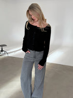 Kvinde ser ned, iført sort trøje og grå jeans, i et minimalistisk rum med lys væg og en sort stol.