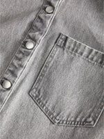 Brystlomme placeret på en grå denimskjorte med sølvfarvede trykknapper. Stoffets tekstur er tydelig i nærbilledet.