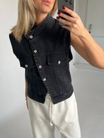 En person tager et spejlbillede iført en sort denimvest og hvide bukser med bindebånd, omgivet af en minimalistisk, lys baggrund.