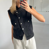 Person tager et spejlbillede iført en sort denimvest og hvide bukser. Personen holder en mobiltelefon med rødmalede negle i et rum med hvide vægge og gulv.