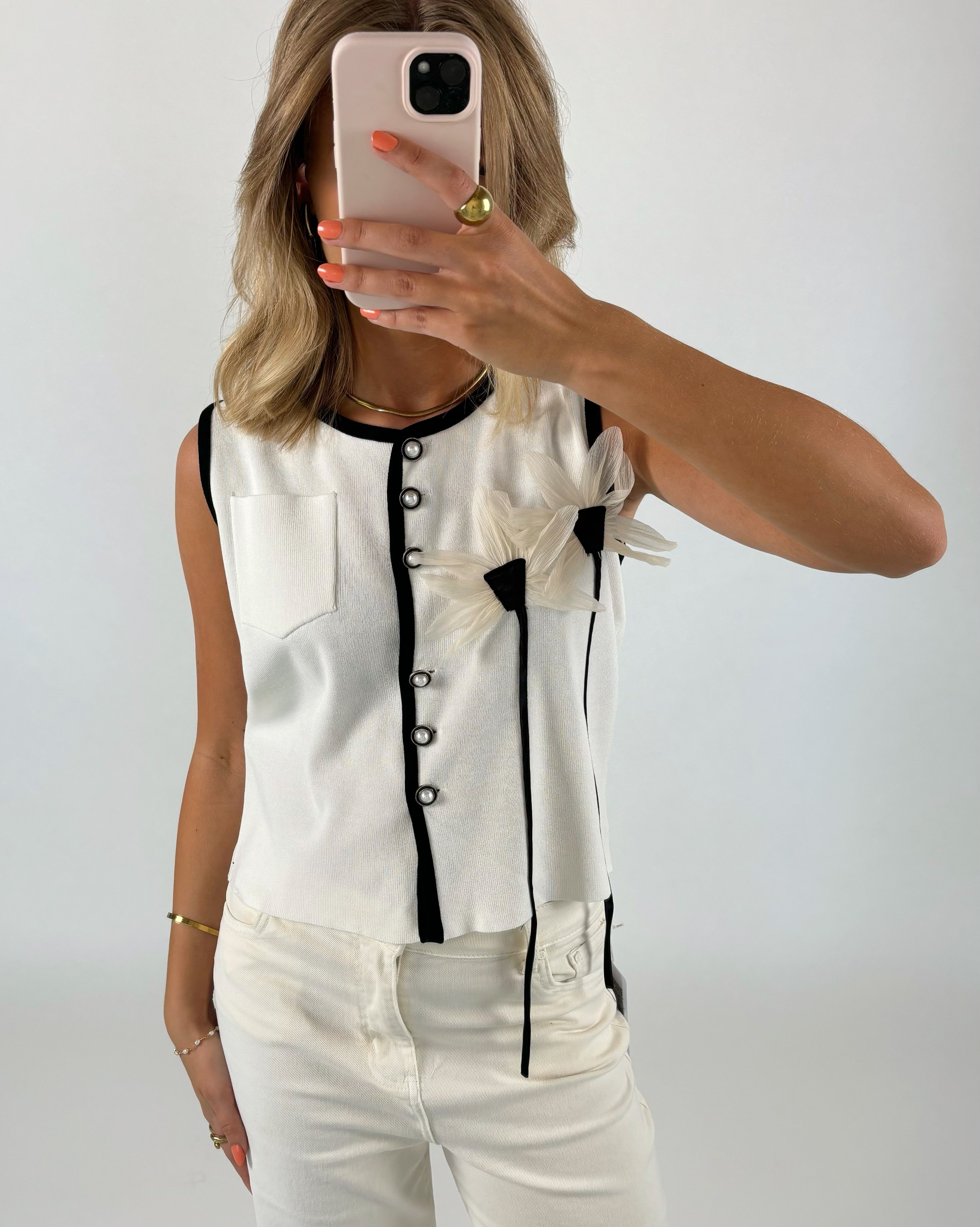 Person tager selfie iført hvid vest med sorte kanter og knapper, dekoreret med blomstermotiver. Hun står i et neutralt rum med mobiltelefonen, iført hvide bukser og orange neglelak.