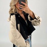 Person tager et spejlbillede iført en sort og beige jakke samt blå jeans. Personen holder en smartphone foran ansigtet i et neutralt baggrundsrum.