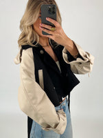 Person tager et spejlbillede iført en sort og beige jakke samt blå jeans. Personen holder en smartphone foran ansigtet i et neutralt baggrundsrum.