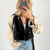 En person tager et spejlbillede iført en sort og beige jakke og blå jeans. Personen holder en telefon op foran ansigtet, omgivet af en lys baggrund.