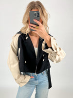 En person tager et spejlbillede iført en sort og beige jakke og blå jeans. Personen holder en telefon op foran ansigtet, omgivet af en lys baggrund.