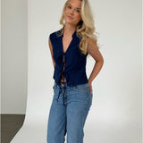 Kvinde står iført en sort, ærmeløs top og blå jeans i en studiolignende baggrund med en lys hvid væg og grå gulv. Hun ser roligt mod kameraet.