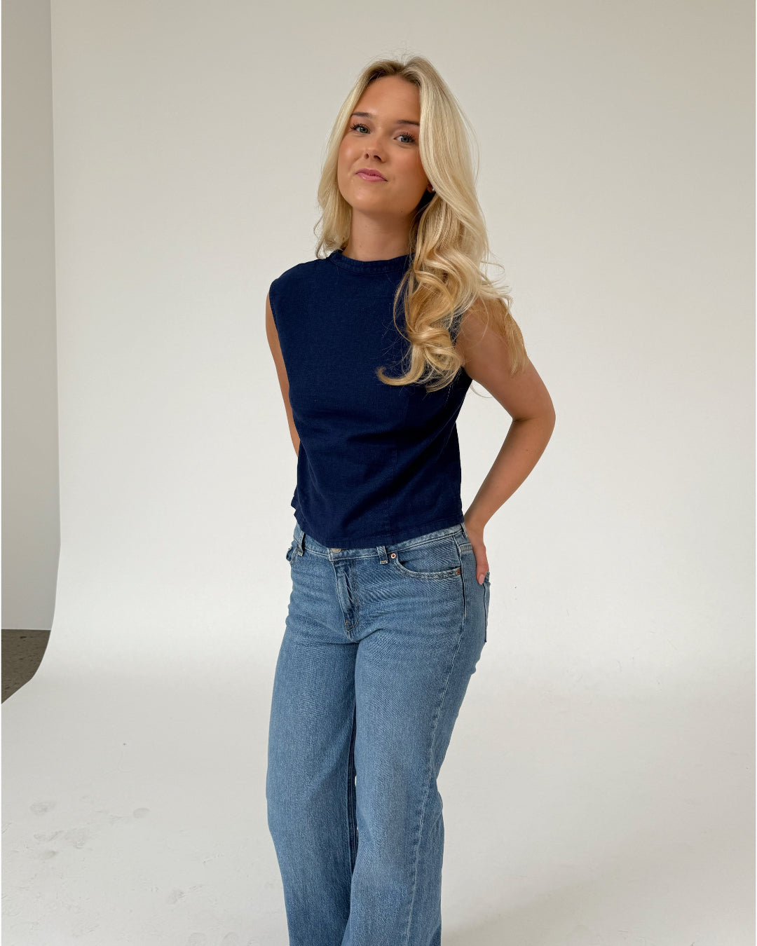 En person står afslappet iført en mørkeblå ærmeløs top og blå jeans i et neutralt studie med en lys baggrund.