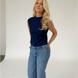 En person står afslappet iført en mørkeblå ærmeløs top og blå jeans i et neutralt studie med en lys baggrund.