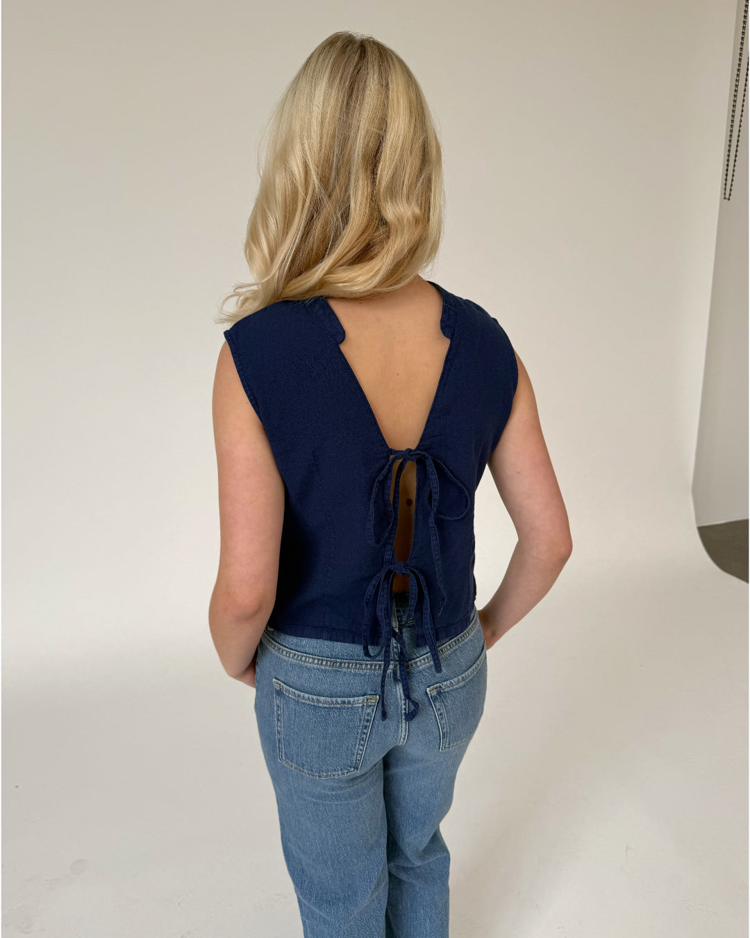 En person med blond hår iført en mørk bluse med åben ryg og bindebånd. Personen bærer jeans og står i et lyst rum uden synlige dekorationer.