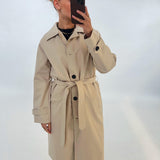 En person iført en beige trenchcoat holder en smartphone foran ansigtet. Baggrunden er ensfarvet hvid, hvilket fremhæver jakken og telefonen.