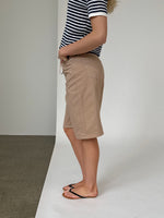 Person iført en stribet trøje og beige shorts står sidevendt, med hænderne i lommerne og iført sorte flip-flops. Baggrunden er enkel med en skillelinje mellem gulv og væg.