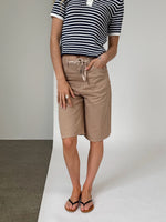 Person i stribet trøje og beige knælange shorts står oprejst med hænderne på siden. Flade sandaler bæres, og baggrunden er en todelt væg, grå og hvid.
