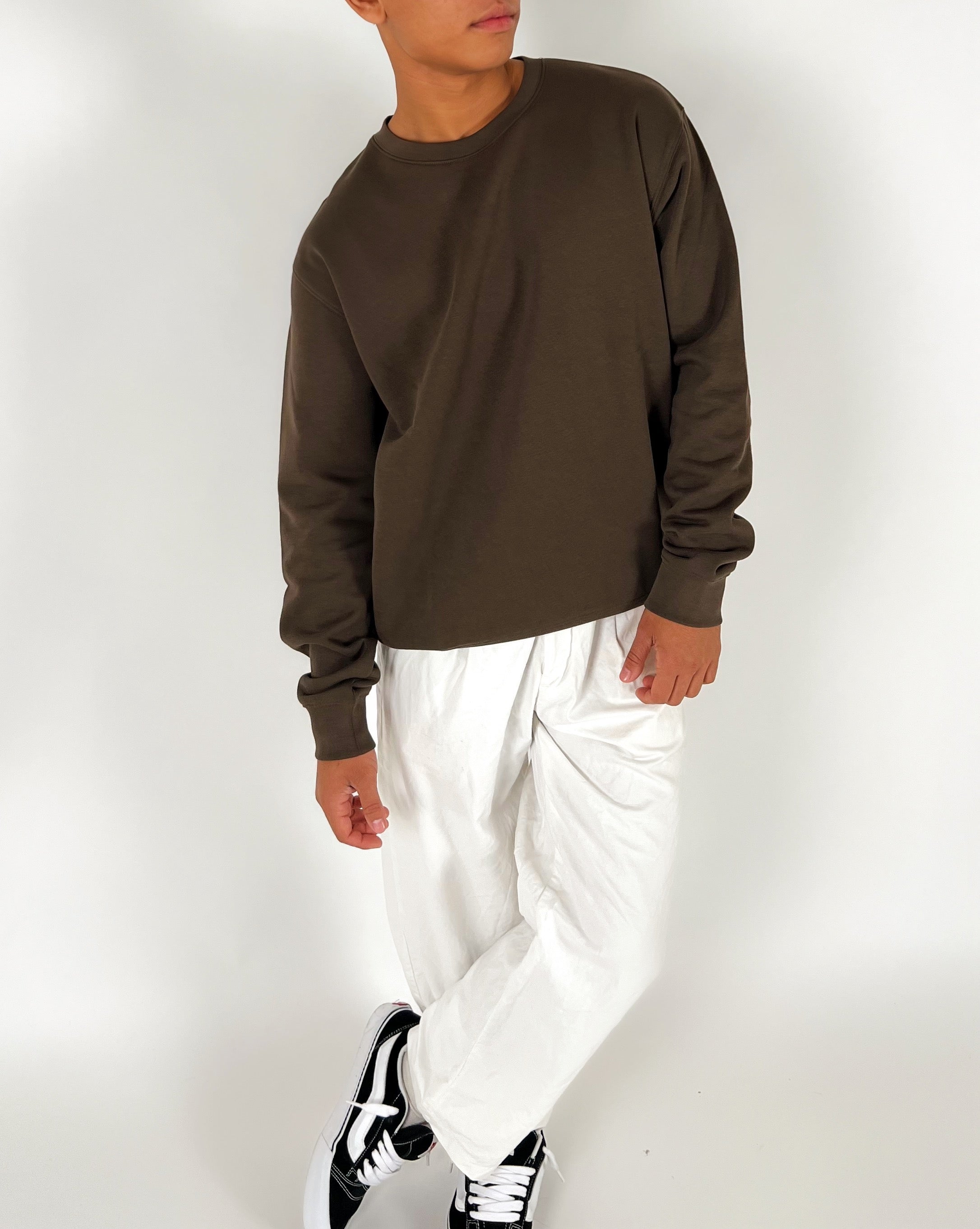 En person iført en brun sweatshirt og hvide bukser står afslappet med krydsede ben i et hvidt rum. Personen bærer sorte og hvide sneakers.