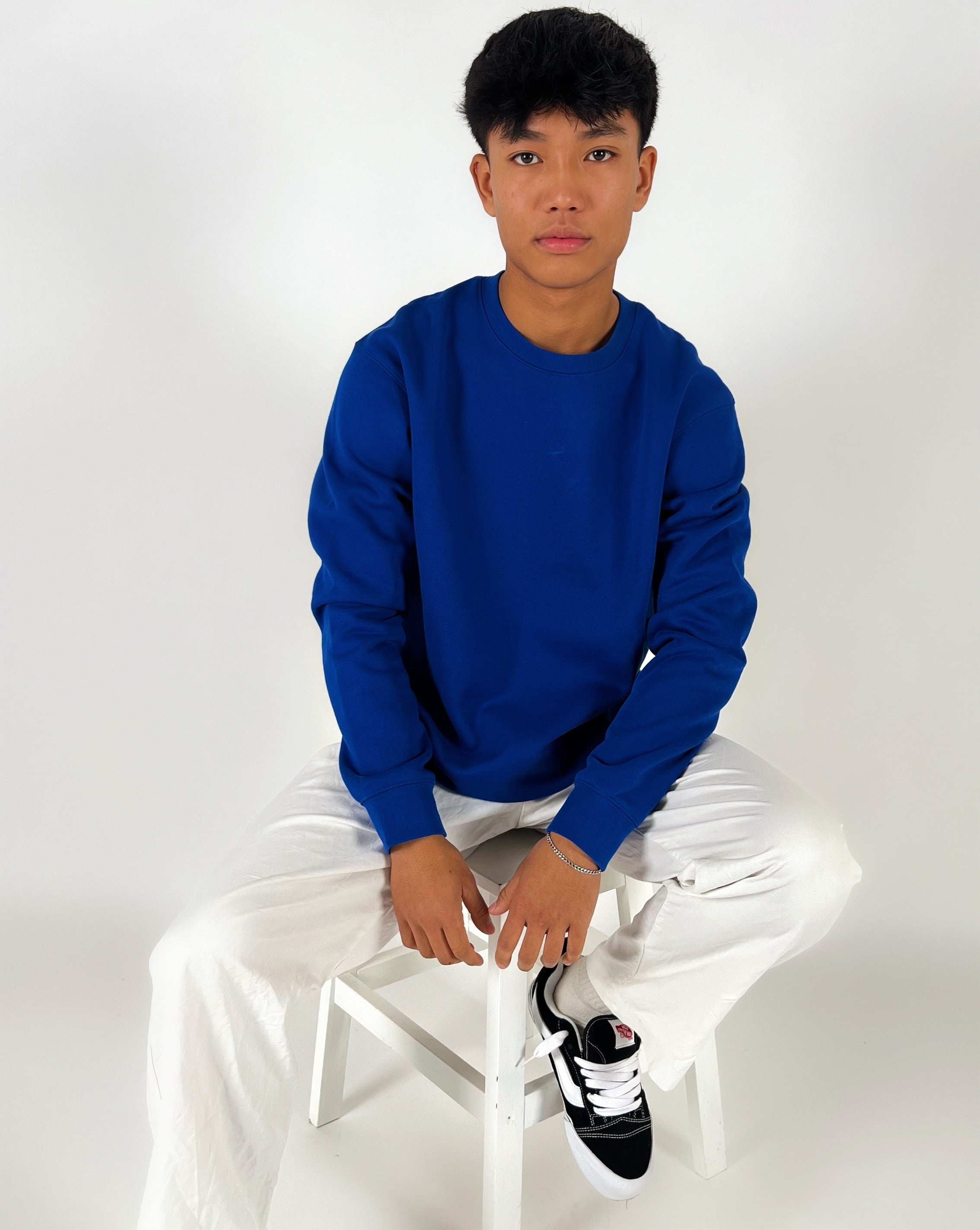 En person sidder på en hvid stol iført en blå sweatshirt og hvide bukser, med sorte og hvide sko, i et neutralt, hvidt rum.