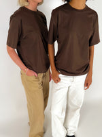 To personer iført brune t-shirts står i et neutralt rum. Den ene har beige bukser, den anden hvide. Begge står afslappet, delvist vendt mod hinanden.