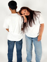 En person står med ryggen til iført en hvid T-shirt og jeans, mens en anden person i lignende tøj hviler hovedet på den første persons skulder, i et neutralt rum.