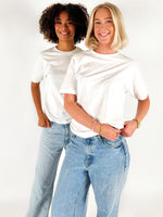 To personer i hvide T-shirts og jeans står smilende i et studie med hvid baggrund. Den ene person har krøllet hår, den anden har skulderlangt blond hår.