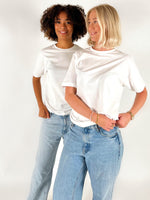 To kvinder iført hvide T-shirts og blå jeans står ved siden af hinanden og smiler i et lyst studiemiljø.