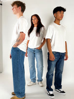 Tre personer står i studie iført hvide T-shirts og blå jeans, med neutral baggrund og lys belysning. To mænd står på hver sin side af kvinden.