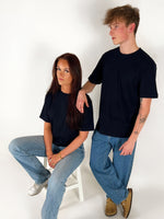 En person sidder på en hvid stol, iført en sort t-shirt og jeans, mens en anden person står ved siden af og hviler en arm på stolens ryglæn. Begge er i et lyst rum.