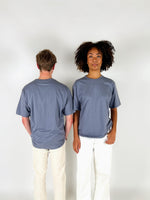 To personer står ved siden af hinanden; en vender ryggen til, mens den anden ser fremad. De bærer begge grå t-shirts og hvide bukser, i et neutralt rum.