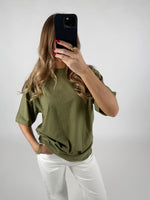 En person iført en grøn T-shirt og hvide bukser holder en sort mobiltelefon foran deres ansigt. Baggrunden er lys og neutral.