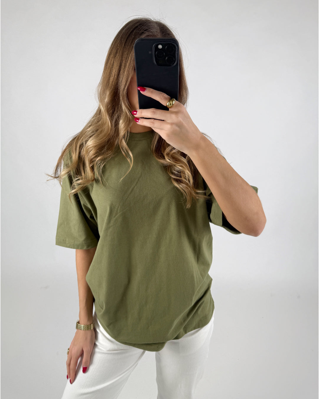 En person iført en olivengrøn T-shirt tager et spejlbillede med en smartphone i hånden. Personen står i et lyst rum med hvid baggrund.