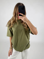 En person iført en olivengrøn T-shirt tager et spejlbillede med en smartphone i hånden. Personen står i et lyst rum med hvid baggrund.