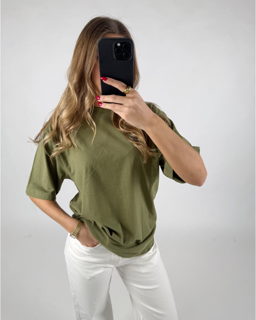 Kvinde tager en selfie med en sort telefon. Hun har en grøn t-shirt og hvide bukser på, i et neutralt oplyst rum. Hendes negle er rødlakkerede.