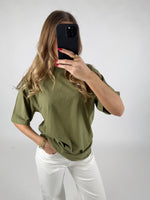 Kvinde tager en selfie med en sort telefon. Hun har en grøn t-shirt og hvide bukser på, i et neutralt oplyst rum. Hendes negle er rødlakkerede.