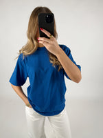 En person iført en blå T-shirt tager et billede med en smartphone foran sit ansigt. Personen har langt hår og står i et neutralt rum.
