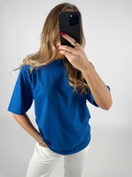 En person i en blå t-shirt holder en smartphone op foran ansigtet i en selfie-position. Personen står i et lyst rum med hvid baggrund.