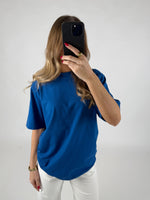 En person med langt hår står i en blå t-shirt og hvide bukser og holder en sort telefon op foran ansigtet i et lysomt rum.