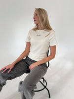 En person sidder på en stol iført en hvid t-shirt med teksten "ASOS DESIGN" og grå jeans. Baggrunden er en simpel, lys væg.