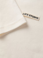 En hvid T-shirt har en påsyet etikette med teksten "JJXX STUDIO" i sorte bogstaver, set i en lys, ensartet baggrund.