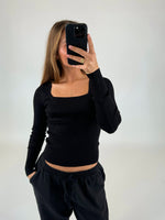 En person iført sort langærmet t-shirt og bukser tager et spejlbillede med en smartphone i en minimalistisk baggrund. Hånden er tæt på kameraet, og hovedet er tildækket.