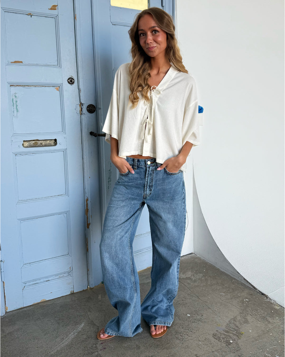 En kvinde står afslappet med hænderne i lommerne, iført jeans og en løs hvid bluse, foran en lyseblå dør i et rum med grå gulv.