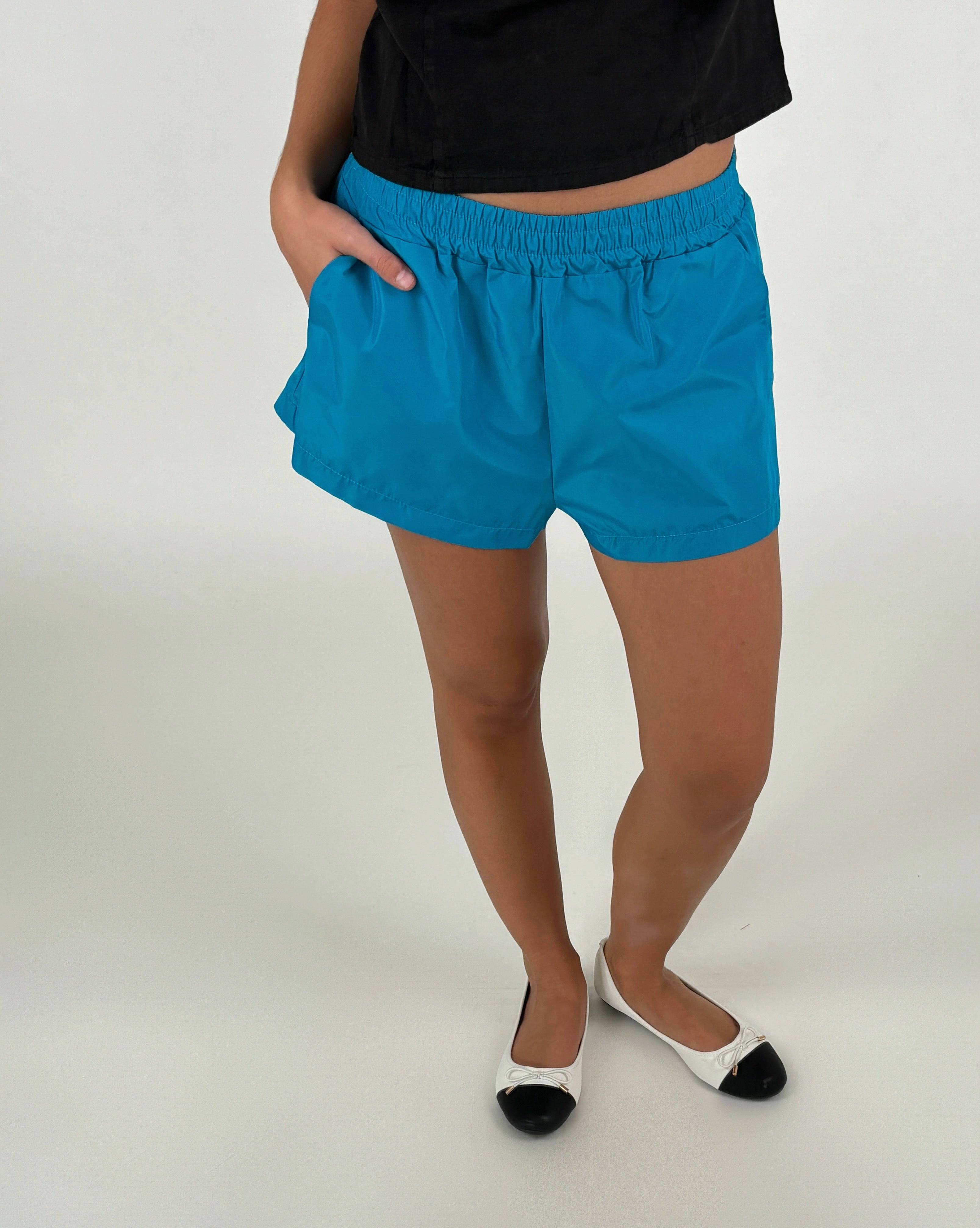 En person står med hånden i lommen på blå shorts, iført sorte og hvide ballerinaer. Personen bærer en sort top og står på et neutralt, lyst underlag.