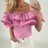 En person iført en pink, off-shoulder top og hvide bukser holder en smartphone og tager et billede i et lyst rum. Personen har blondt hår og bærer guldsmykker.