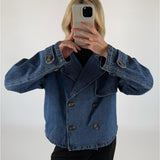 En person i en dobbeltknappet denimjakke tager et spejlselfie med en smartphone i lyse omgivelser. Personen har sort tøj under jakken.