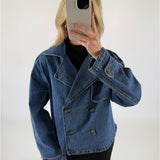 Person i blå denimjakke tager et spejlbillede med en smartphone, står i et rum med en hvid baggrund.