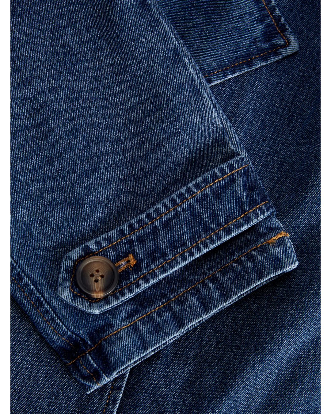 En blå denimjakke har en knap fastgjort på ærmet. Stoffet er detaljeret med orange syninger, der skaber et klassisk og robust look.