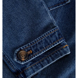 En blå denimjakke har en knap fastgjort på ærmet. Stoffet er detaljeret med orange syninger, der skaber et klassisk og robust look.